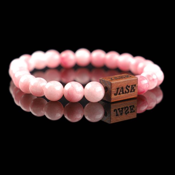 Handmade pink agate beaded bracelet with a dark wood engraved 'JASE' logo block, shown on a reflective black surface.