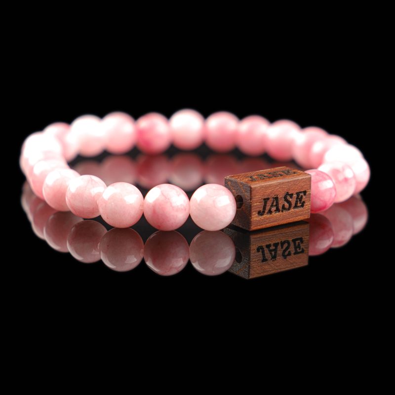 Handmade pink agate beaded bracelet with a dark wood engraved 'JASE' logo block, shown on a reflective black surface.