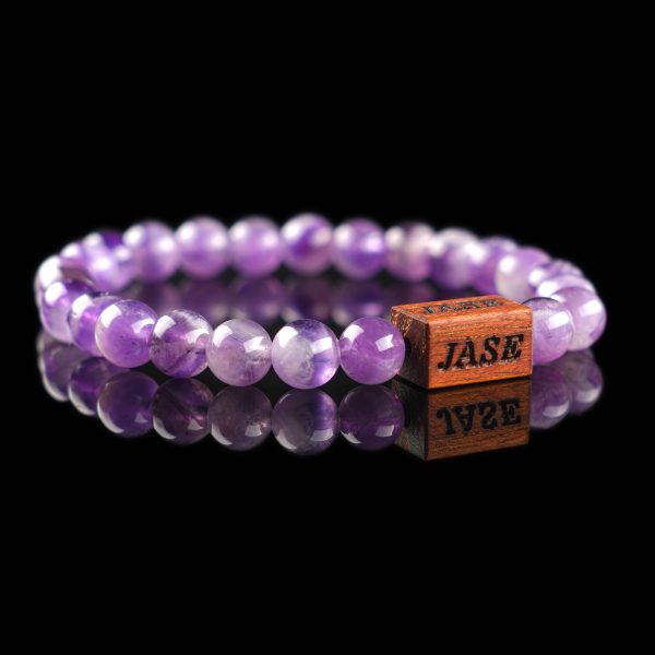 Handmade purple Amethyst beaded bracelet with a dark wood engraved 'JASE' logo block, shown on a reflective black surface.