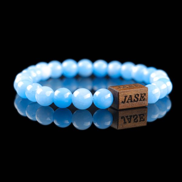 Handmade light blue aquamarine beaded bracelet with a dark wood engraved 'JASE' logo block, shown on a reflective black surface.
