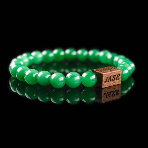 Handmade green aventurine beaded bracelet with a dark wood engraved 'JASE' logo block, shown on a reflective black surface.