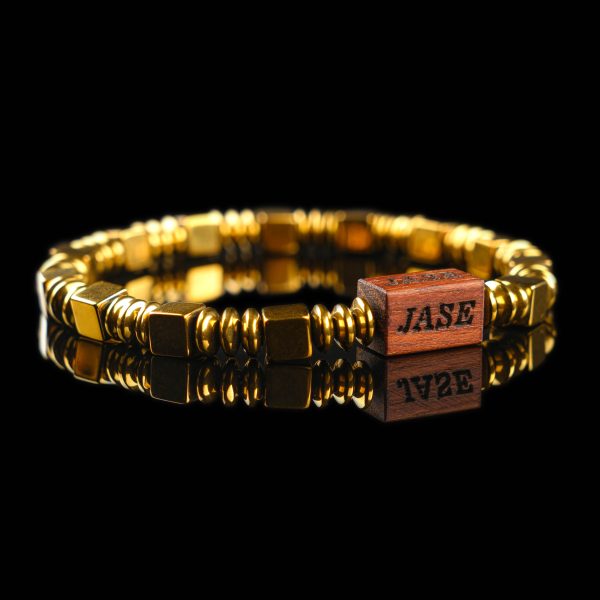 Handmade golden hematite cube bead bracelet with a dark wood engraved 'JASE' logo block, shown on a reflective black surface.