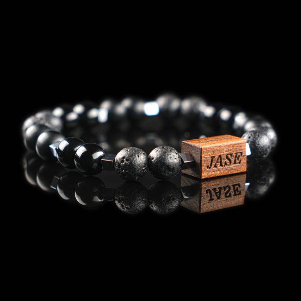 Handmade bracelet with black lava, matte onyx, and Grey hematite cube beads, featuring a dark wood engraved 'JASE' logo block on a reflective black surface.