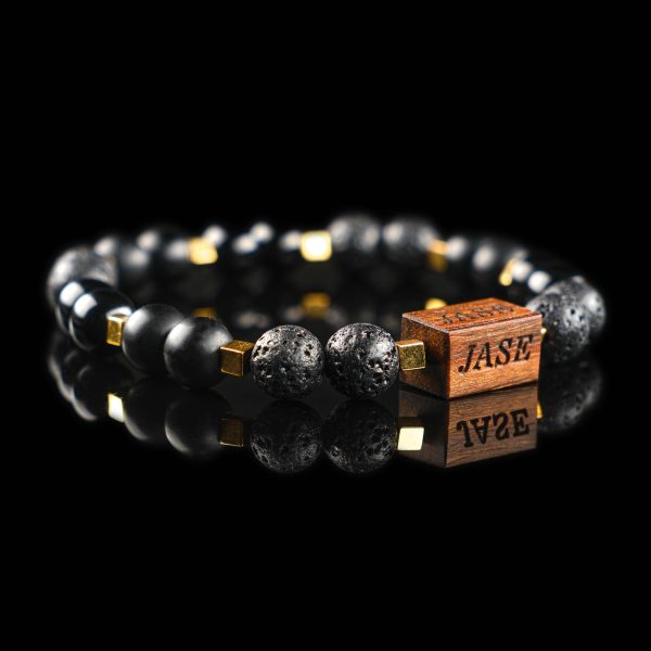 Handmade bracelet with black lava, matte onyx, and golden hematite cube beads, featuring a dark wood engraved 'JASE' logo block on a reflective black surface.