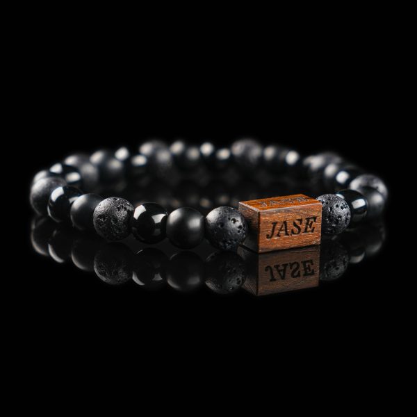 Handmade bracelet with black lava stone, polished onyx, and matte onyx beads, featuring a dark wood engraved 'JASE' logo block on a reflective black surface.