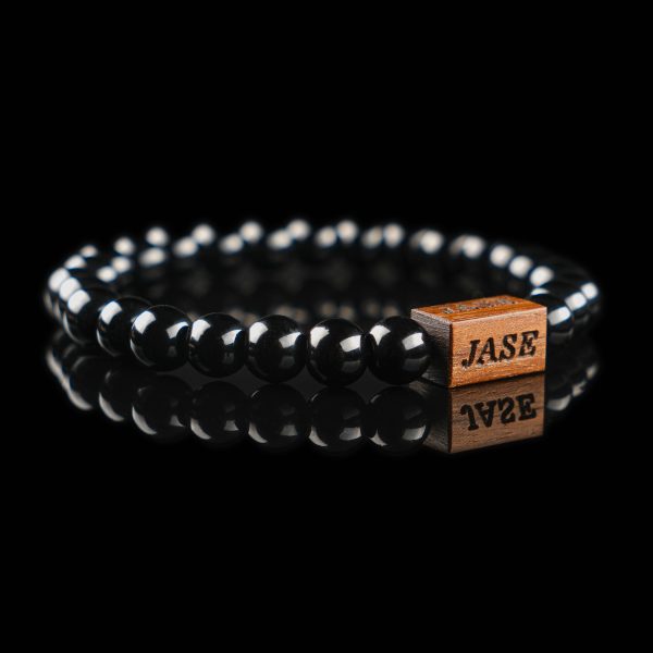 Handmade polished black onyx beaded bracelet with a dark wood engraved 'JASE' logo block, shown on a reflective black surface.