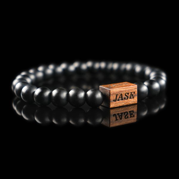 Handmade polished Grey onyx beaded bracelet with a dark wood engraved 'JASE' logo block, shown on a reflective black surface.