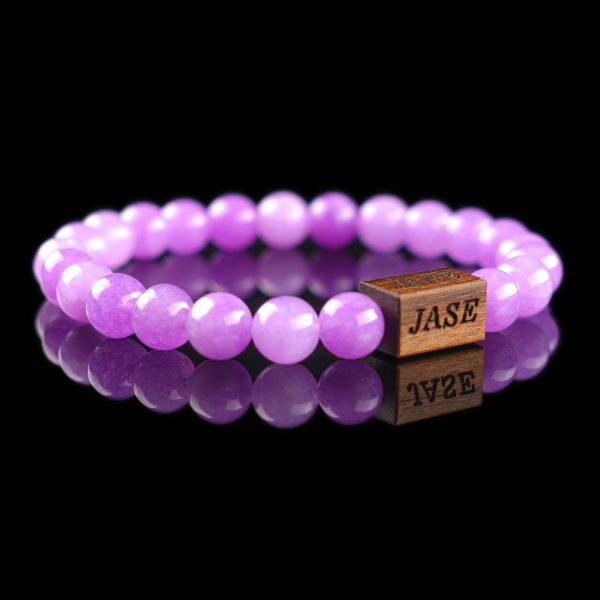 Handmade lilac jade beaded bracelet with light purple stones and a dark wood engraved 'JASE' logo block, shown on a reflective black surface.