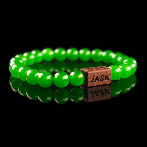 Handmade green jade beaded bracelet with vibrant green stones and a dark wood engraved 'JASE' logo block, shown on a reflective black surface.