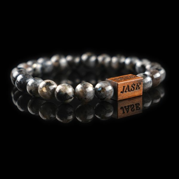 Handmade labradorite beaded bracelet with grey and black stones showing a subtle flash of labradorescence, with a dark wood engraved 'JASE' logo block, on a reflective black surface.