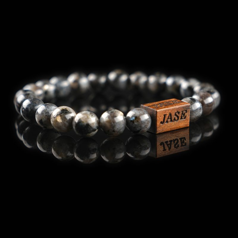 Handmade labradorite beaded bracelet with grey and black stones showing a subtle flash of labradorescence, with a dark wood engraved 'JASE' logo block, on a reflective black surface.