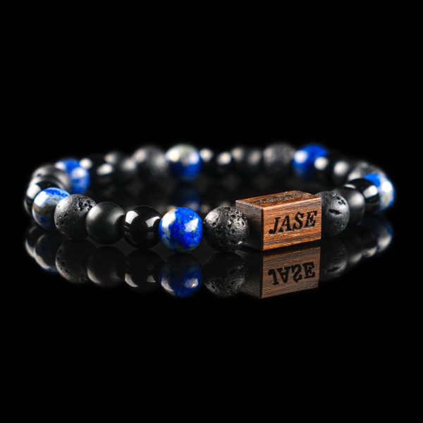 Handmade bracelet with black lava stone, matte onyx, and deep blue lazurite beads, featuring a dark wood engraved 'JASE' logo block on a reflective black surface.