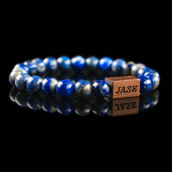Handmade deep blue lazurite beaded bracelet with golden pyrite flecks and a dark wood engraved 'JASE' logo block, shown on a reflective black surface.