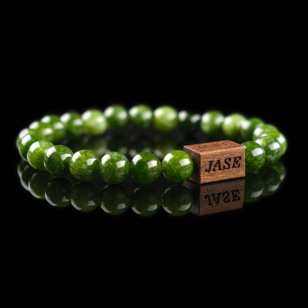 Handmade nephrite jade beaded bracelet with deep green stones and a dark wood engraved 'JASE' logo block, shown on a reflective black surface.