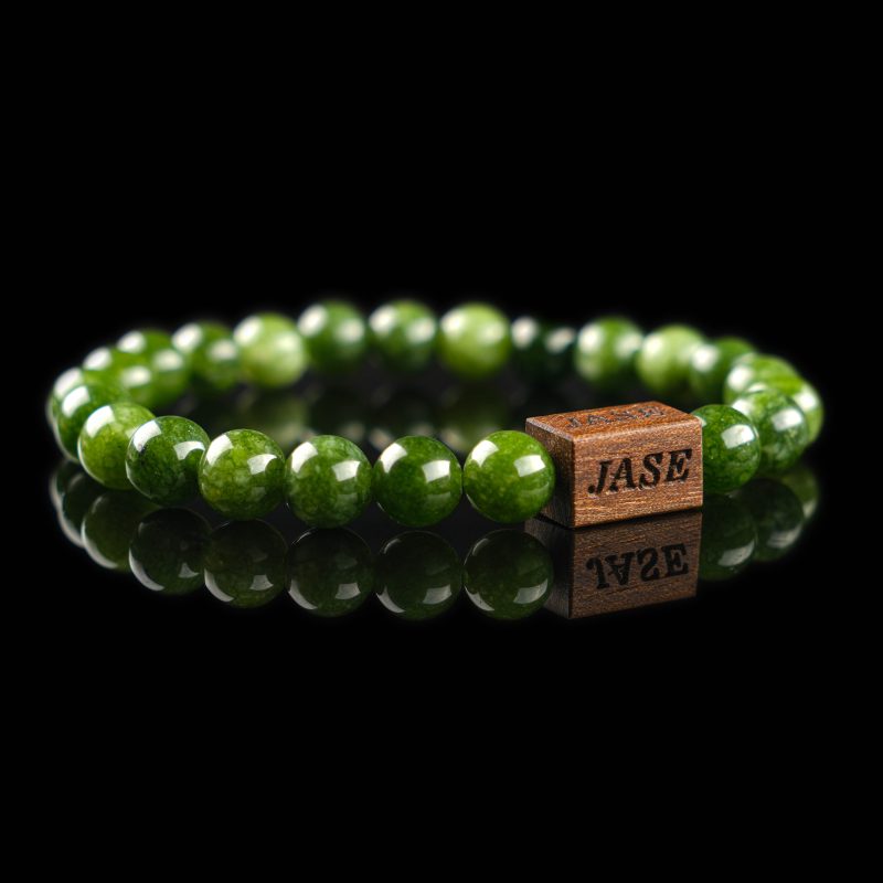 Handmade nephrite jade beaded bracelet with deep green stones and a dark wood engraved 'JASE' logo block, shown on a reflective black surface.
