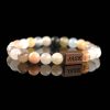 Handmade multi-color quartz beaded bracelet with soft pastel stones in peach, white, and yellow, and a dark wood engraved 'JASE' logo block, shown on a reflective black surface.