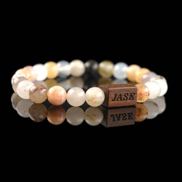 Handmade multi-color quartz beaded bracelet with soft pastel stones in peach, white, and yellow, and a dark wood engraved 'JASE' logo block, shown on a reflective black surface.