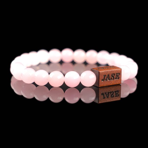 Handmade faceted rose quartz beaded bracelet with soft pink stones and a dark wood engraved 'JASE' logo block, shown on a reflective black surface.