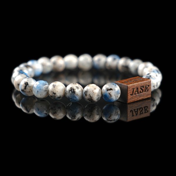 Handmade sodalite beaded bracelet with blue and black spots on a light grey stone matrix, featuring a dark wood engraved 'JASE' logo block on a reflective black surface.