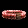 Handmade strawberry quartz beaded bracelet with vibrant pinkish-red stones and a dark wood engraved 'JASE' logo block, shown on a reflective black surface.