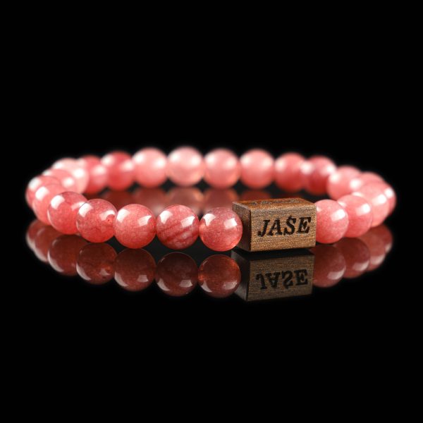 Handmade strawberry quartz beaded bracelet with vibrant pinkish-red stones and a dark wood engraved 'JASE' logo block, shown on a reflective black surface.