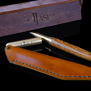 The Legacy handcrafted wood and brass pen set by 1jase, featuring a tan leather case and an engraved wooden gift box on a black reflective surface.